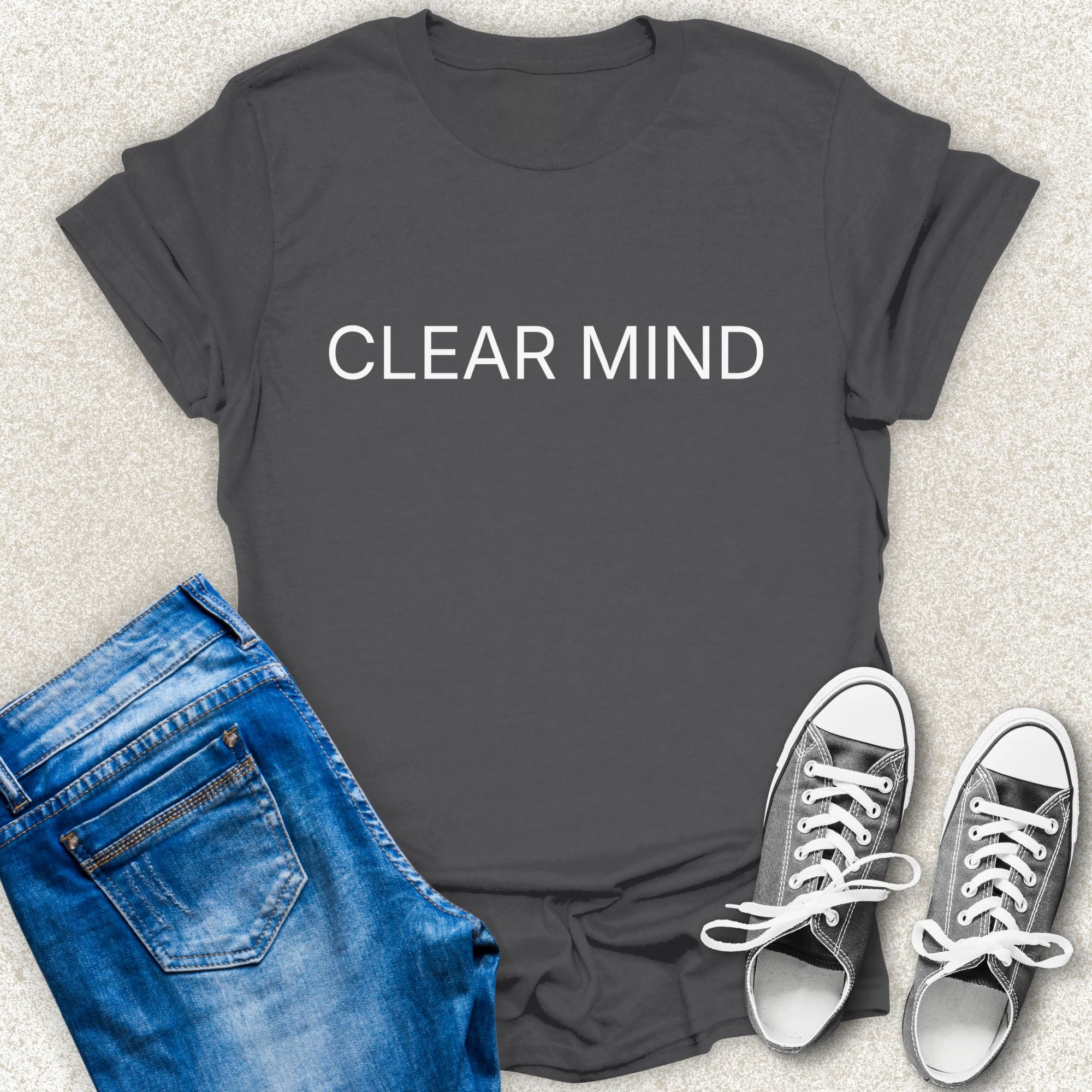 A Charcoal short-sleeve T-shirt with the words 'CLEAR MIND' printed in white across the chest, styled in a flat lay with blue jeans and silver sneakers on a light textured background by Low Signal.
