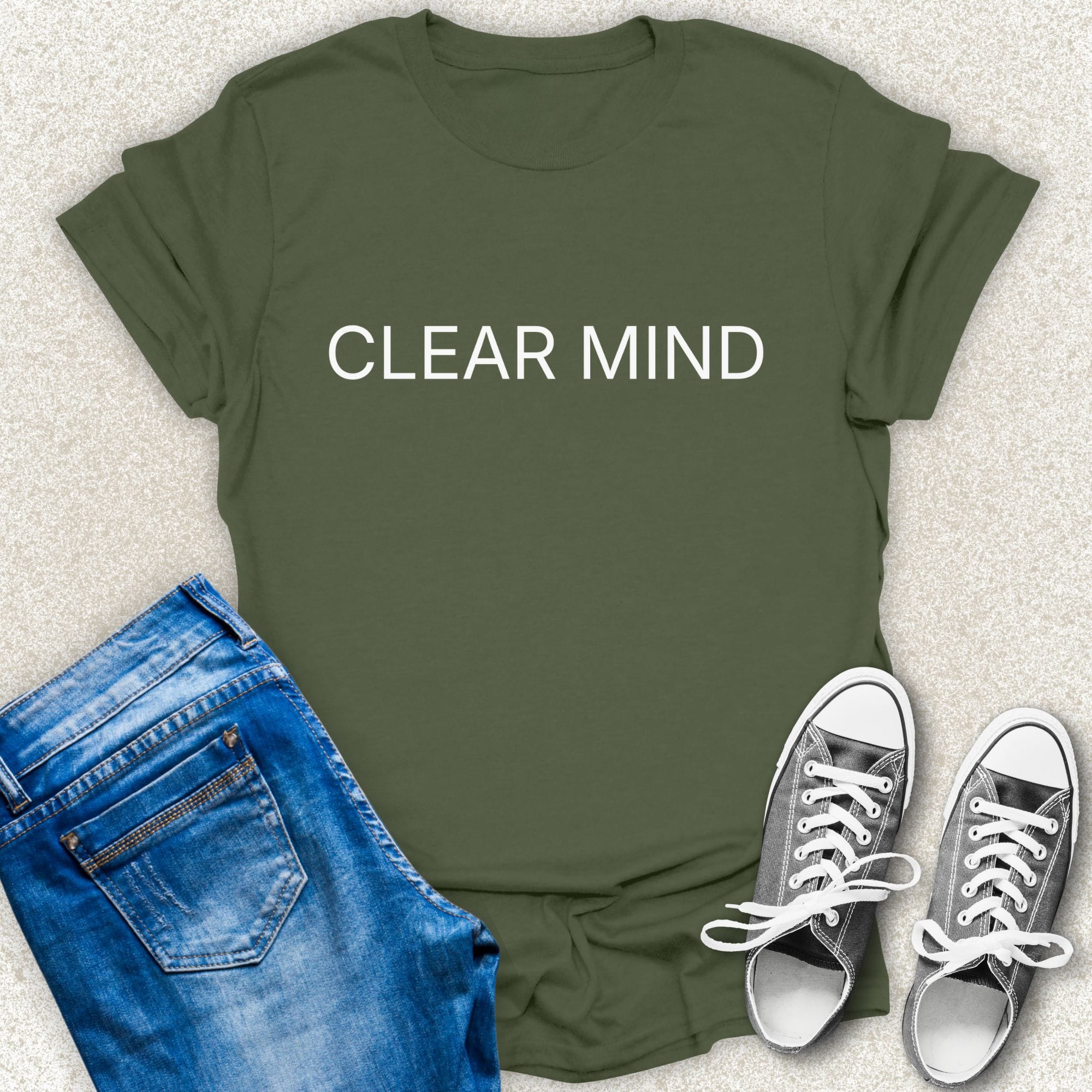 A Military Green short-sleeve T-shirt with the words 'CLEAR MIND' printed in white across the chest, styled in a flat lay with blue jeans and silver sneakers on a light textured background by Low Signal.
