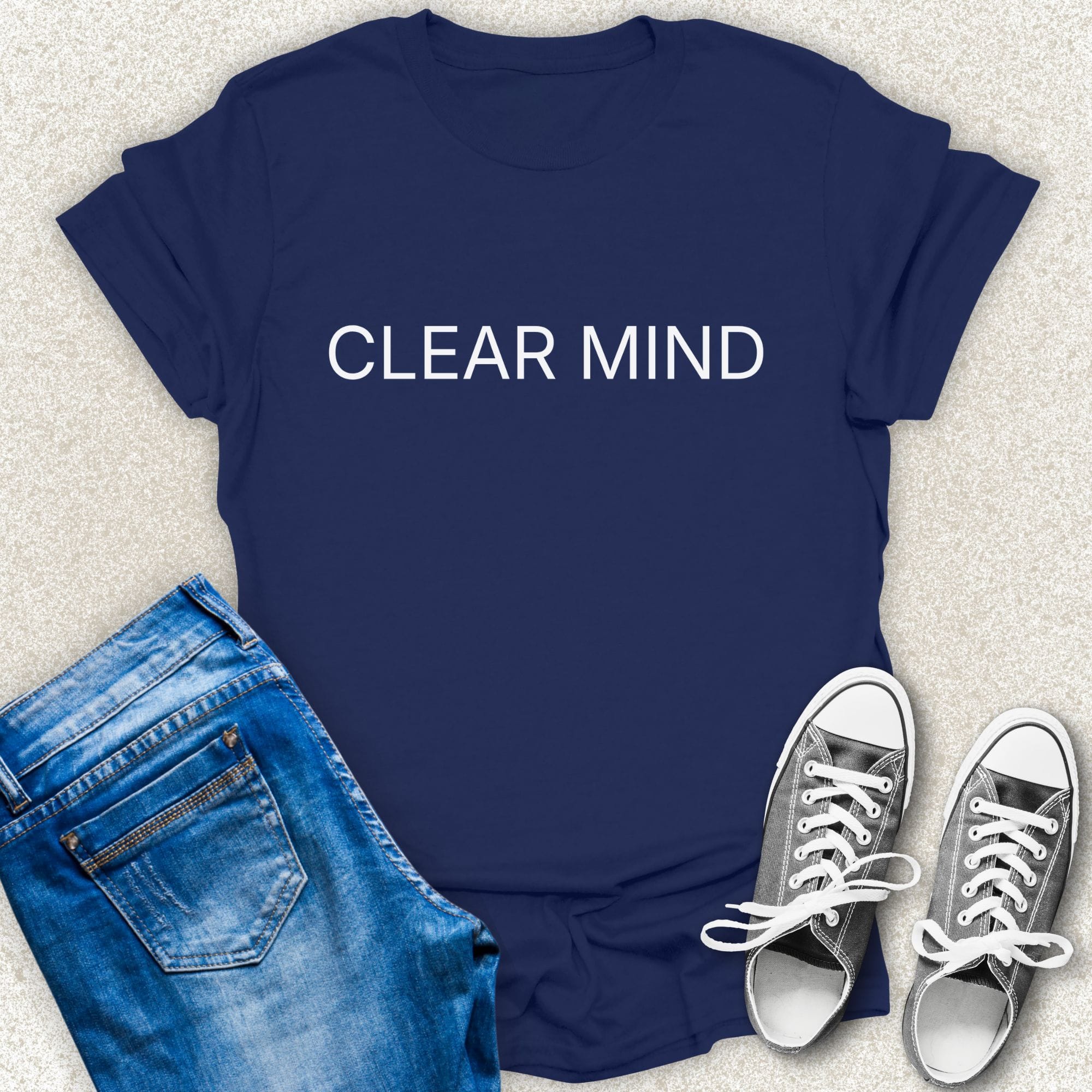 A Navy short-sleeve T-shirt with the words 'CLEAR MIND' printed in white across the chest, styled in a flat lay with blue jeans and silver sneakers on a light textured background by Low Signal.