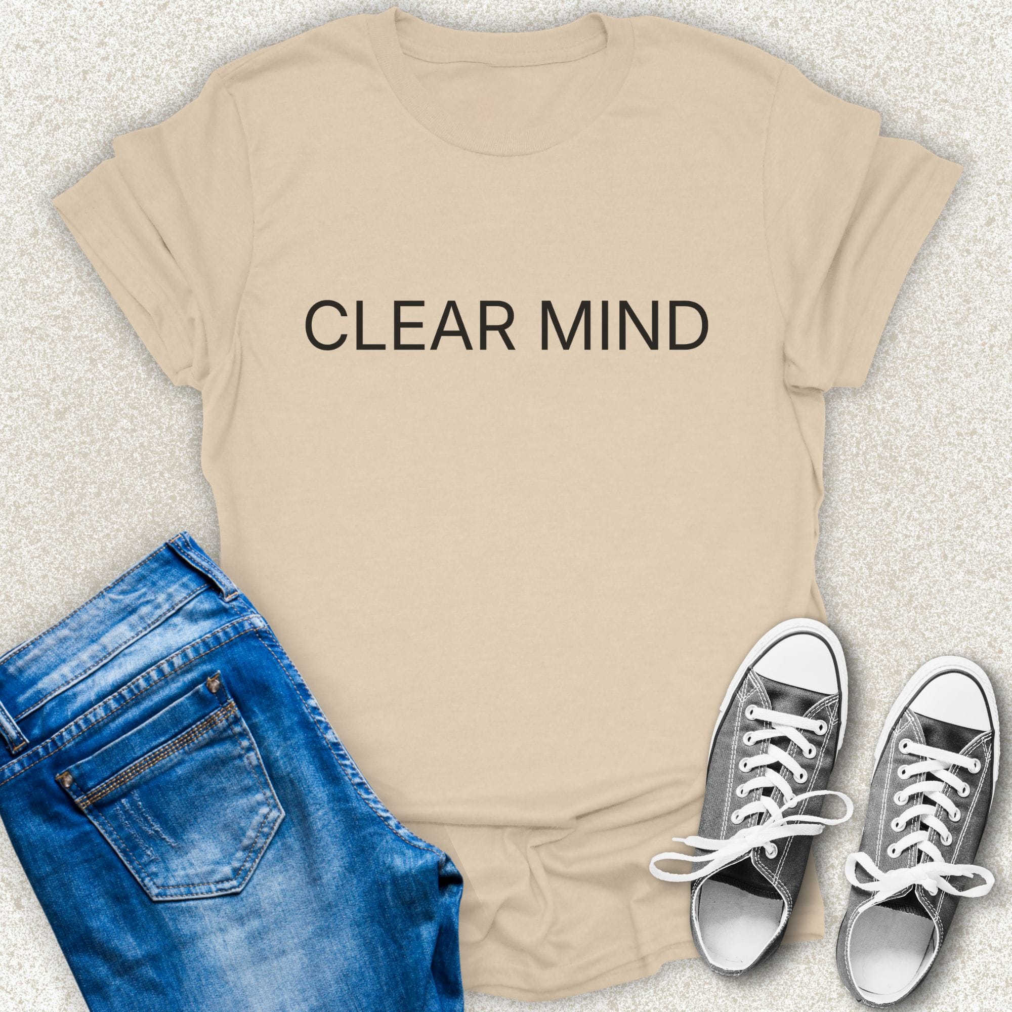 A Sand short-sleeve T-shirt with the words 'CLEAR MIND' printed in white across the chest, styled in a flat lay with blue jeans and silver sneakers on a light textured background by Low Signal.
